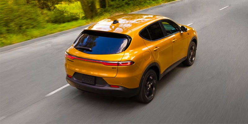 Rear/top view of a glossy yellow SUV driving on a winding paved road with blurred green trees in the background.