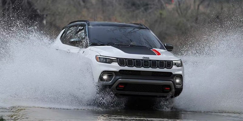 White Jeep SUV with black roof, hood decal, and red stripe, splashing through a shallow stream.
