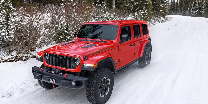 A red Jeep Wrangler Rubicon driving on a snowy road with evergreen trees in the background.