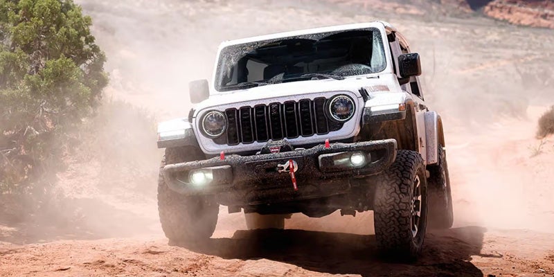 A white Jeep Rubicon 4x4 kicks up dust as it drives forward on a dirt trail.